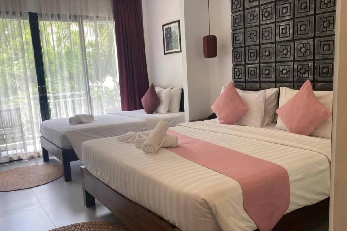A hotel room with two beds, each with a pillow and a rolled towel. There is a window with curtains, a wall with a geometric pattern, and a small pendant light.