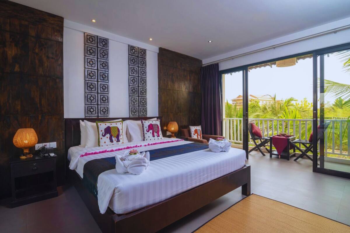 A hotel room with a large bed, a nightstand, a lamp, a balcony with chairs, and a view of palm trees.