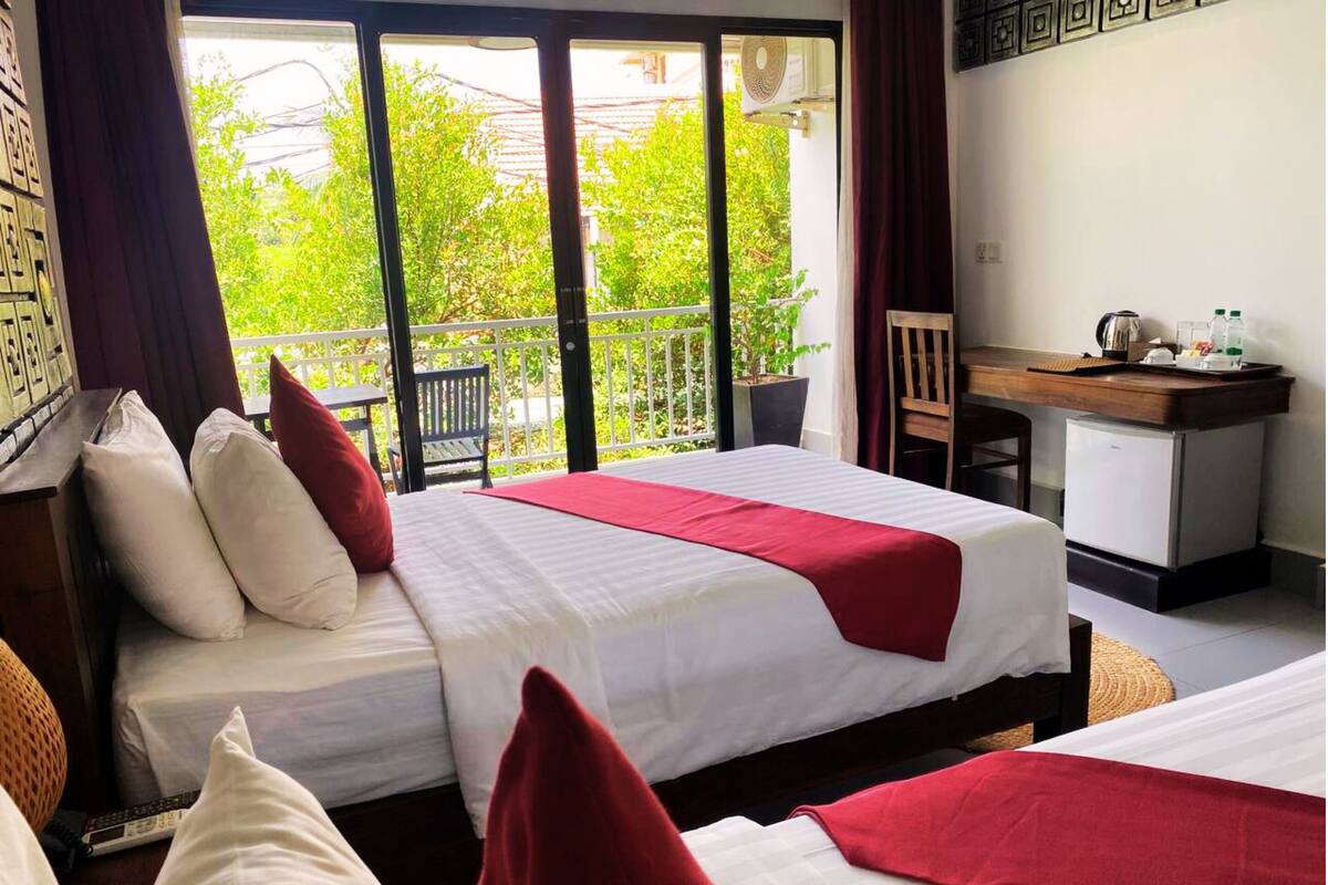 A hotel room with two beds, a desk, and a view of greenery outside.
