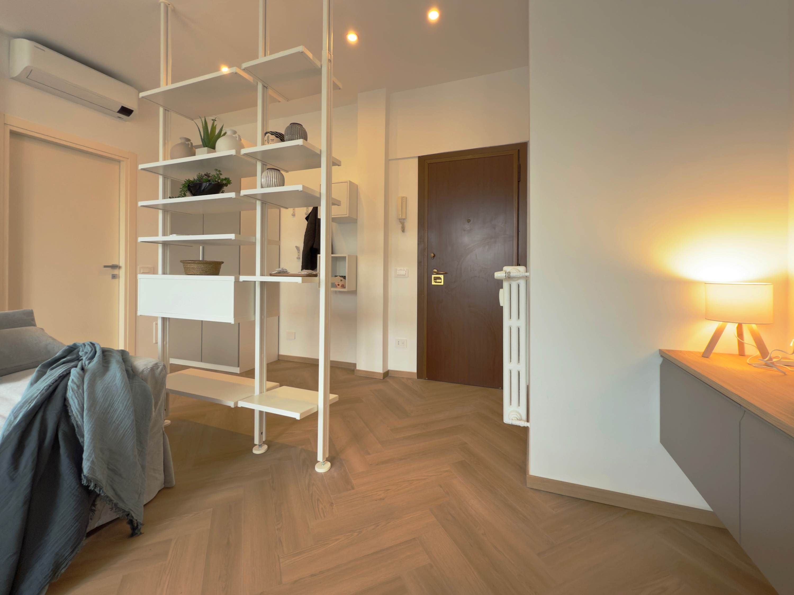 A modern room with a white shelving unit, a bed with a grey blanket, a radiator, and a desk with a lamp.