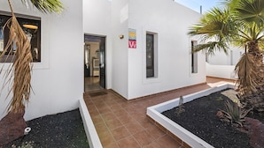 Um edifício moderno com fachada branca, entrada revestida com azulejos e um pequeno jardim com plantas e uma palmeira.