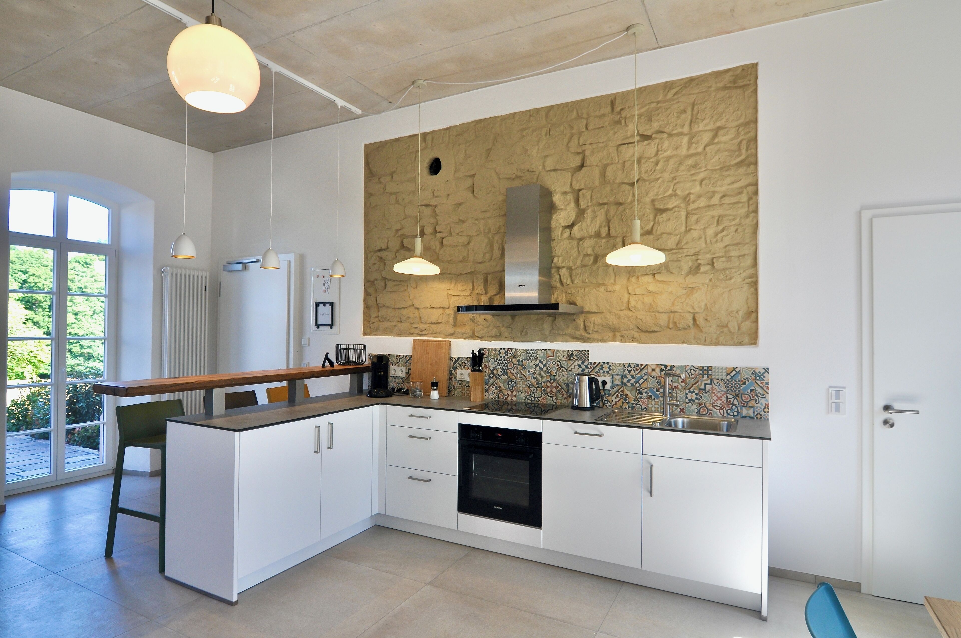 A modern kitchen with white cabinets, a built-in oven, a stone backsplash, pendant lighting, and a view of greenery outside.