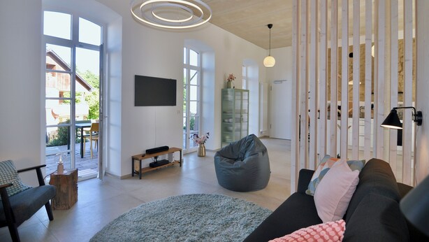 A spacious living room with a large window, a black sofa, a bean bag, and a flat-screen TV mounted on the wall.