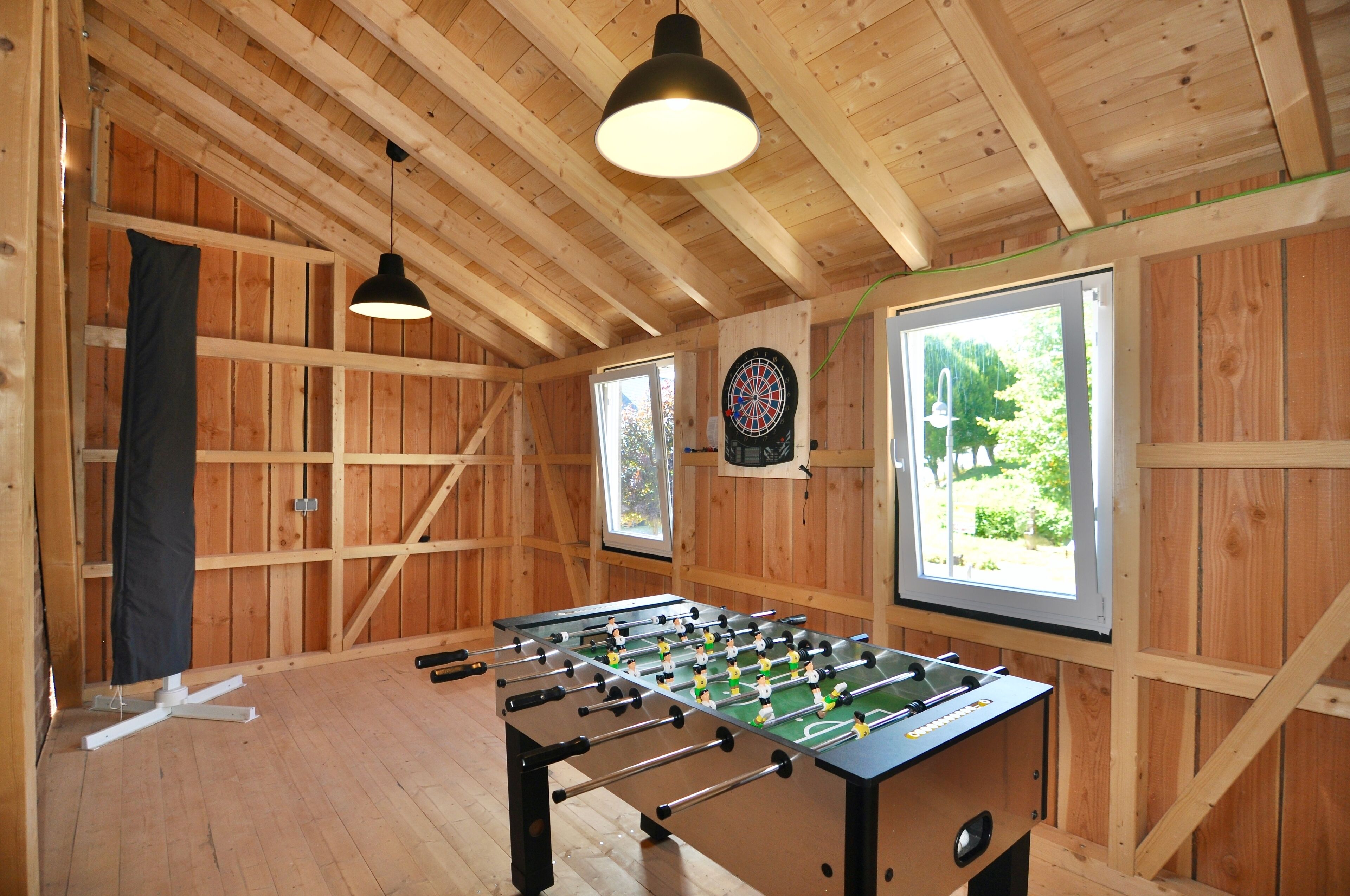 Una habitación con paredes de madera, mesa de fútbol de mesa, dardo y ventana con vista a follaje.