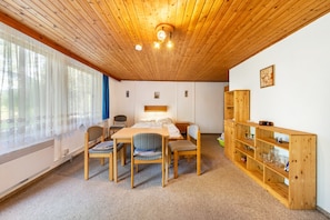 Une pièce avec un plafond en bois, un lit, une table avec des chaises, une bibliothèque et une fenêtre ornée de rideaux légers.
