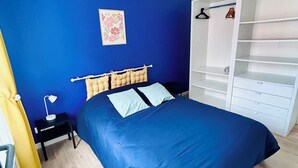 Quarto com parede azul, cama com colcha azul, mesa de cabeceira com abajur, guarda-roupa com prateleiras e um cabideiro.