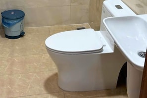 A white toilet with a lid, a blue trash bin, and a white sink in a bathroom.