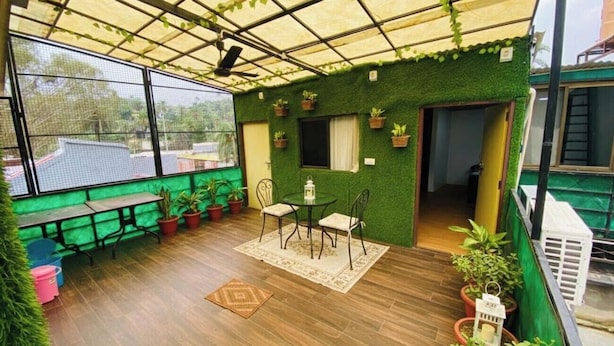 Un espace extérieur abrité doté d’un plancher en bois, d’une table avec des chaises, de plantes en pot et d’une vue sur les bâtiments environnants.