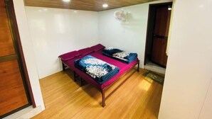 Une petite pièce avec un plafond en bois, un lit recouvert d’une couverture rouge et bleue, et un ventilateur fixé au mur.