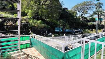 A covered outdoor area with a patterned floor, a green fence, and a view of trees and parked cars in the background.