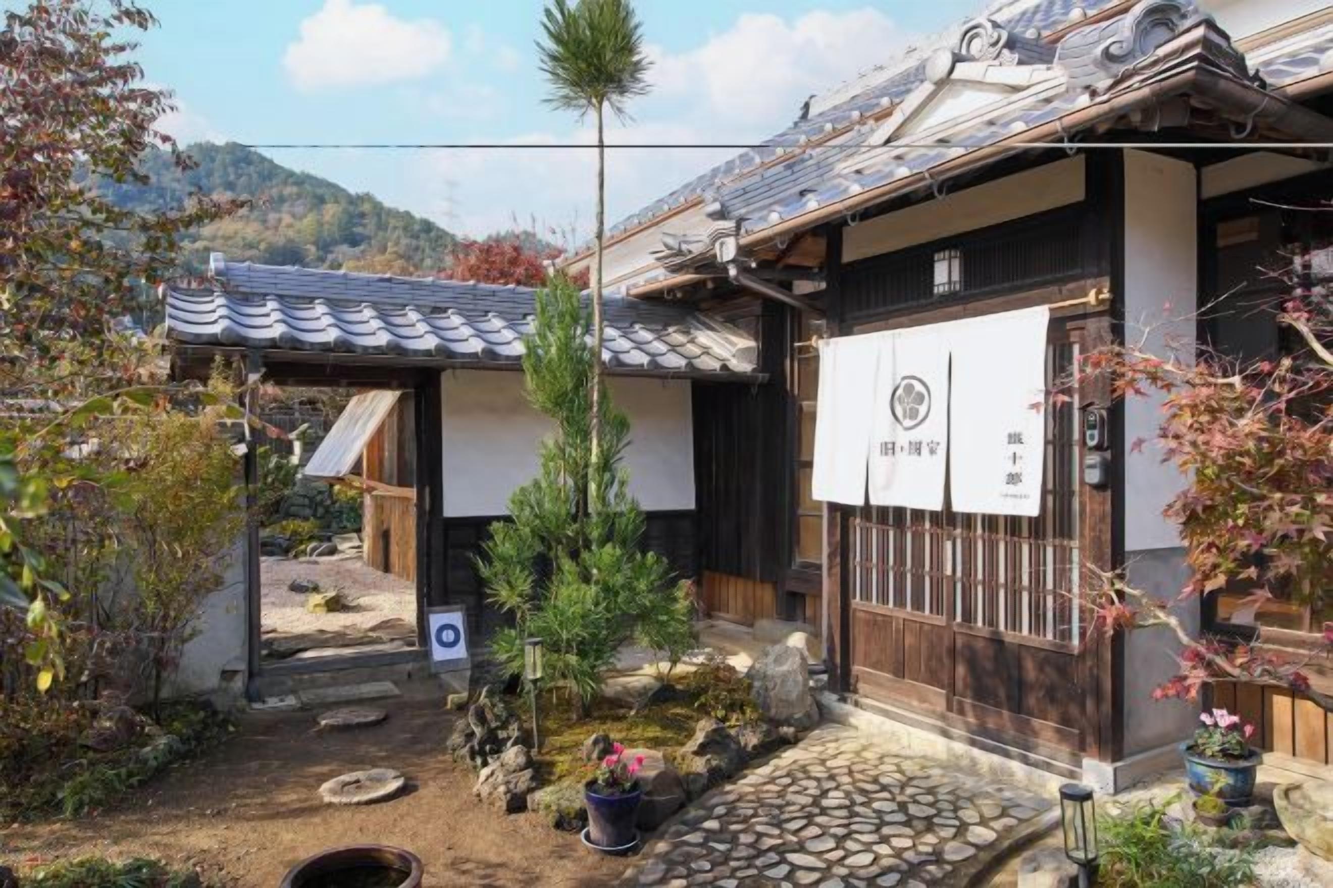 A traditional Japanese building with a tiled roof, a wooden gate, and a small garden with a pine tree.