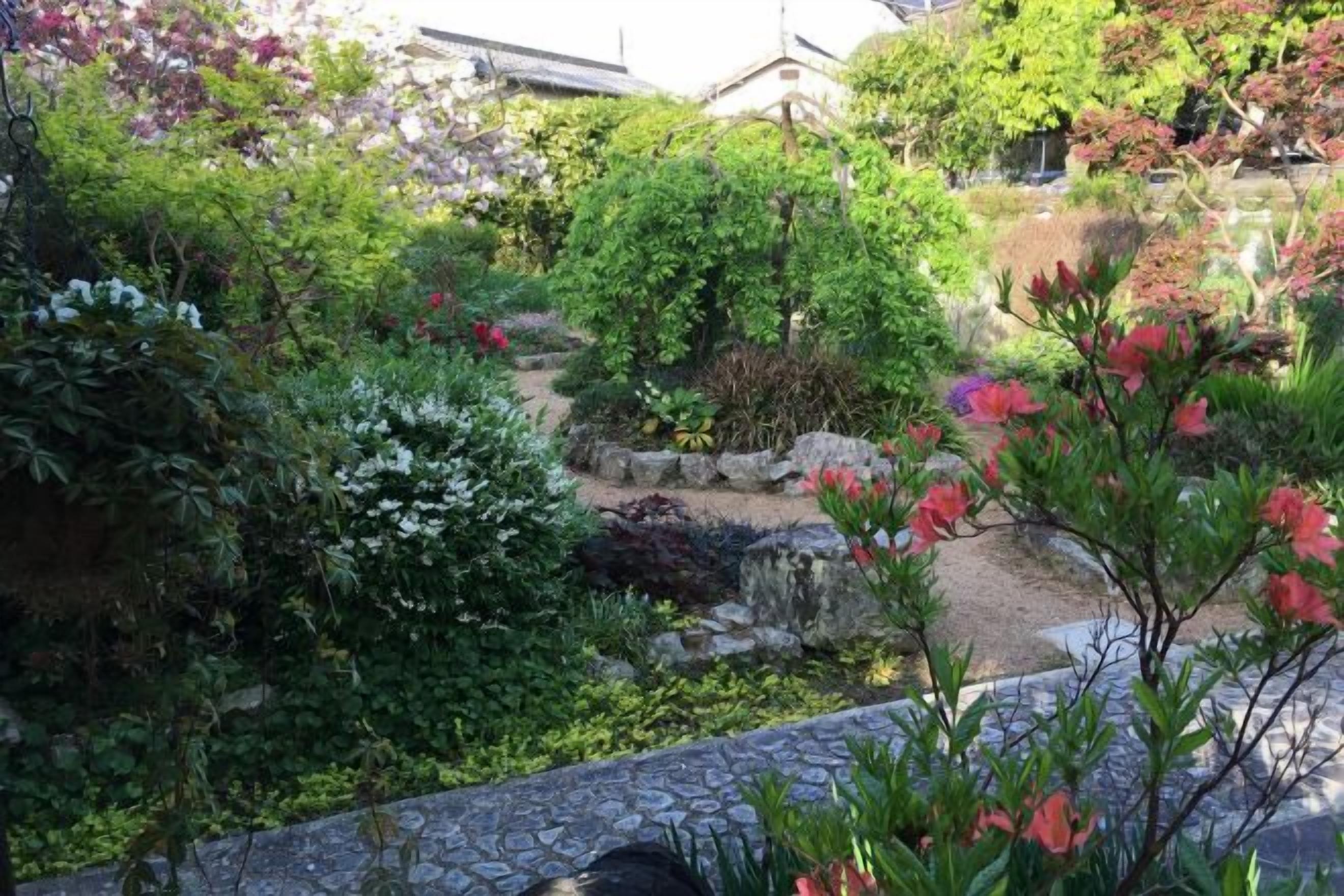Un jardí amb un camí de pedra, diverses plantes i un edifici tradicional al fons.