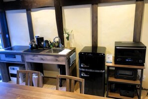 Eine Küchenzeile mit Holztisch, Stühlen, einer Spüle und einer Kaffeemaschine. Zudem befinden sich schwarze Geräte auf einem Regal.