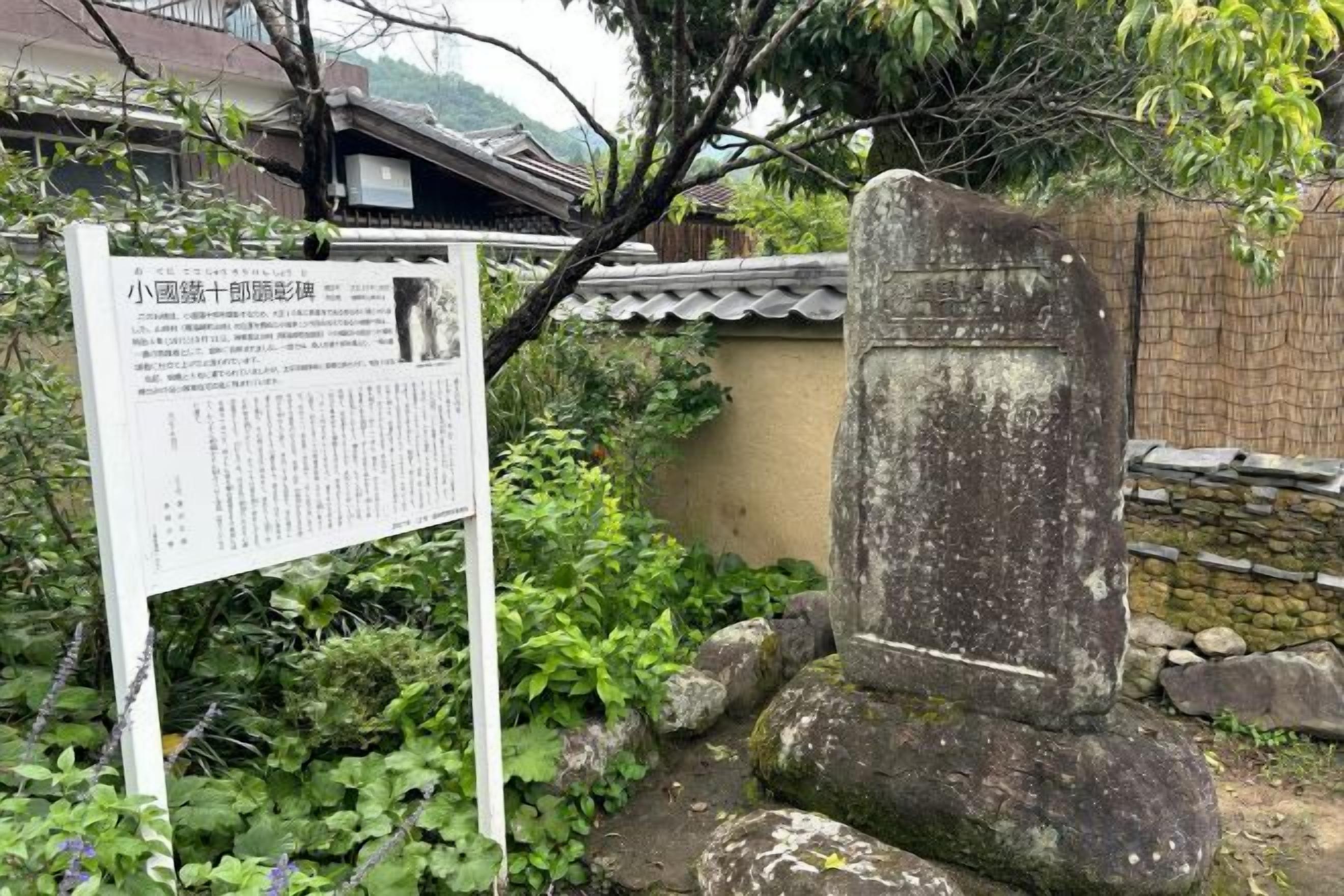 A stone monument with a sign in front of it, surrounded by greenery and a traditional building in the background.