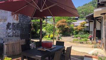 Zona pranzo all'aperto con tavolo e sedie, un ombrellone rosso e vista su un giardino e edifici sullo sfondo.