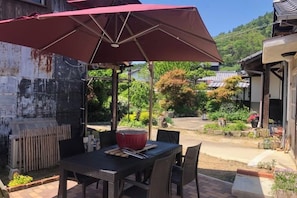 Un espace de restauration en plein air avec une table et des chaises, un parasol rouge, et une vue sur un jardin et des bâtiments en arrière-plan.