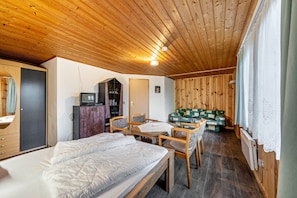 Une pièce aux murs lambrés, comprenant un lit, une table avec des chaises, un buffet et une fenêtre avec des rideaux.