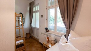 A bedroom with a bed, a wooden ladder, a radiator, a window with curtains, and a small table with a bottle and a lamp.