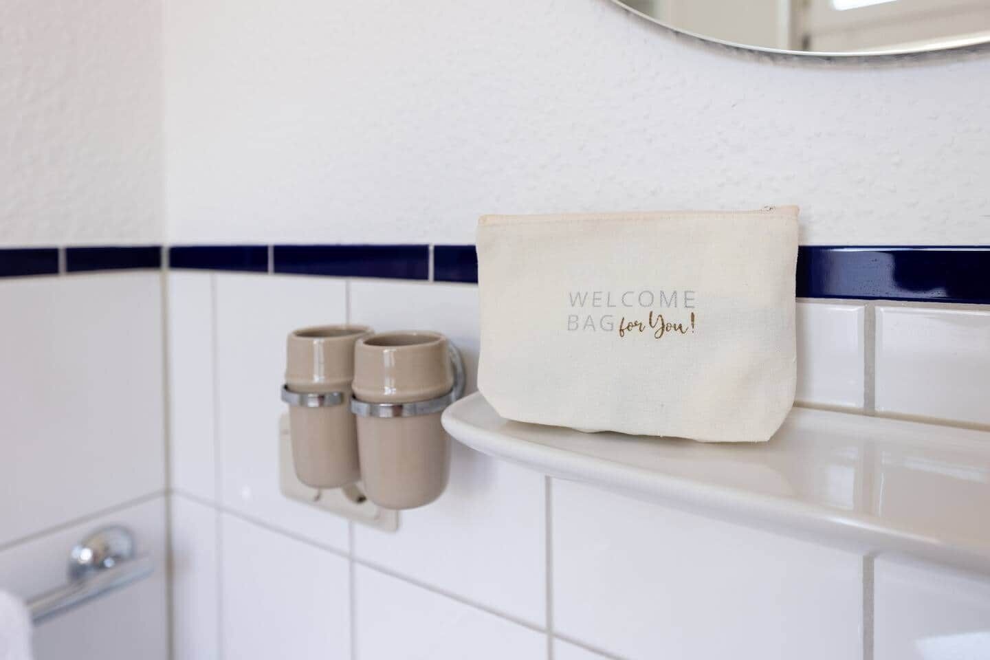Un bagno con un lavandino bianco, due dispenser color beige e una borsa di benvenuto sul ripiano.