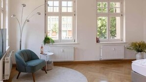 Ein geräumiges Zimmer mit Holzboden, einem runden weißen Teppich, einem türkisfarbenen Sessel, einem kleinen weißen Tisch und zwei Fenstern mit weißen Rahmen.