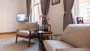 Un salon comprenant un canapé, deux fauteuils, une petite table avec une bouteille et des verres, un téléviseur et une fenêtre avec des rideaux.