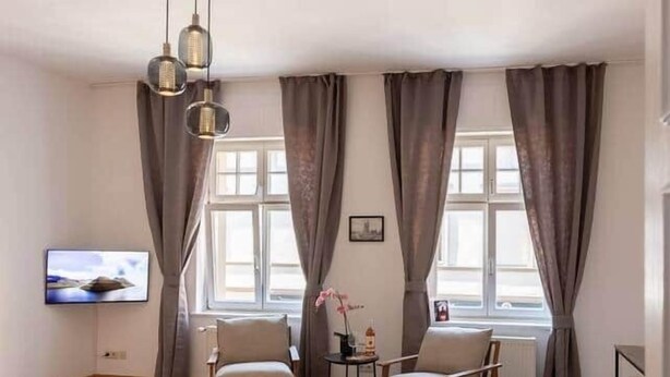 Un salon spacieux doté d’un parquet en bois, de deux fauteuils, d’une petite table, d’un téléviseur fixé au mur et d’une fenêtre avec des rideaux.