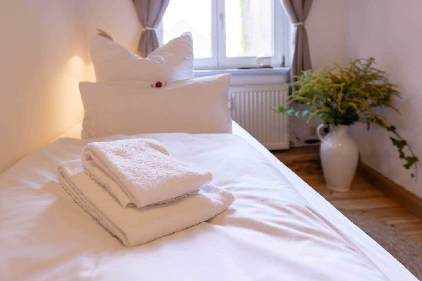 A neatly made bed with white linens and a folded towel, a white pillow, and a vase with a plant on the side.