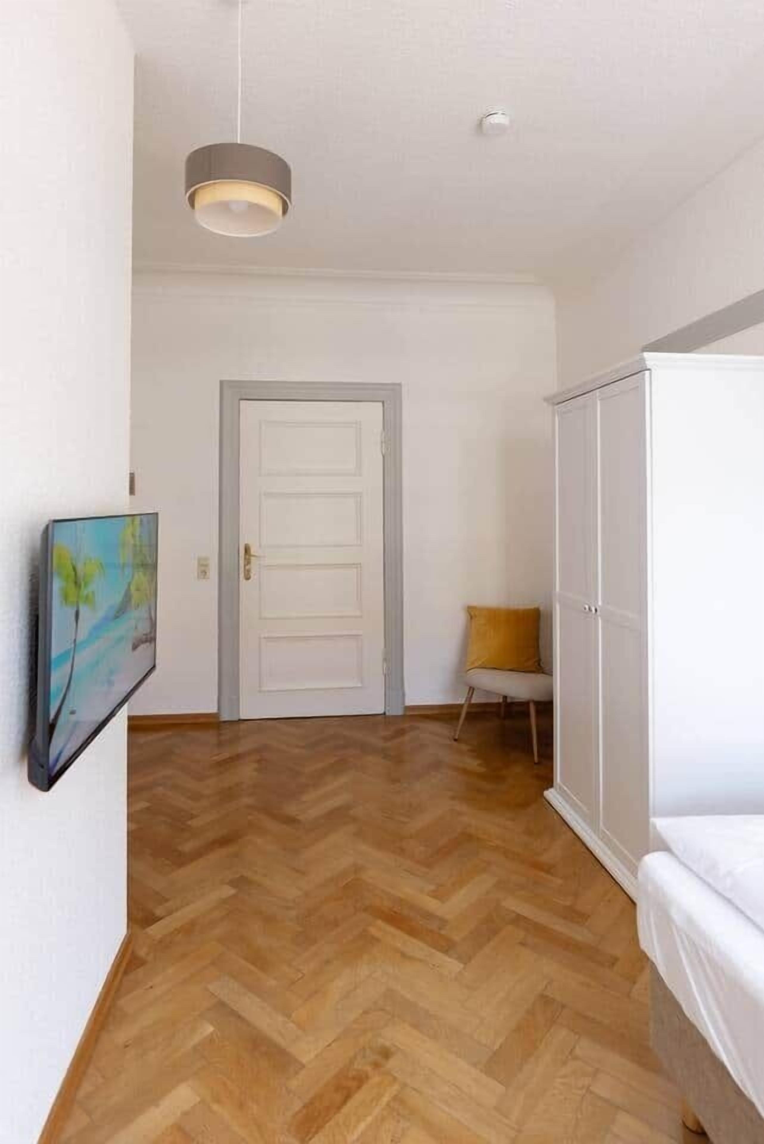 A hallway with a wooden herringbone floor, a white door, a white wardrobe, and a small chair with a yellow cushion.