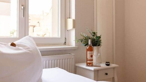 A bedroom with a bed, a window, a bedside table with a bottle and a plant, and a mirror. - Twinbed (Stendal)