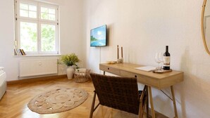 A room with a wooden floor, a dining table with a bottle of wine, a TV on the wall, and a window with a view of greenery. - Cozy Guesthouse at Winckel (Stendal)