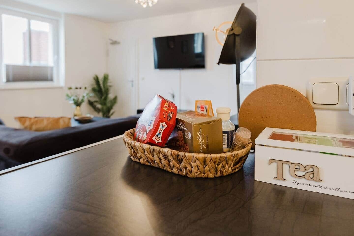 Ein modernes Wohnzimmer mit einem dunklen Holztisch, einem gewebten Korb voller Snacks, einer mit „Tee“ beschrifteten Schachtel und einem fest montierten Flachbildfernseher.