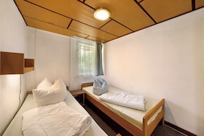 Ein kleines Hotelzimmer mit einem Bett, einem Fenster mit Vorhängen, einer Holzbalkendecke und einem Wandregal.