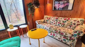 A room with a wooden paneled wall, a floral sofa, a yellow table, a green chair, and a window with a view of trees. - 4 Star Holiday Home in Ry (Ry)