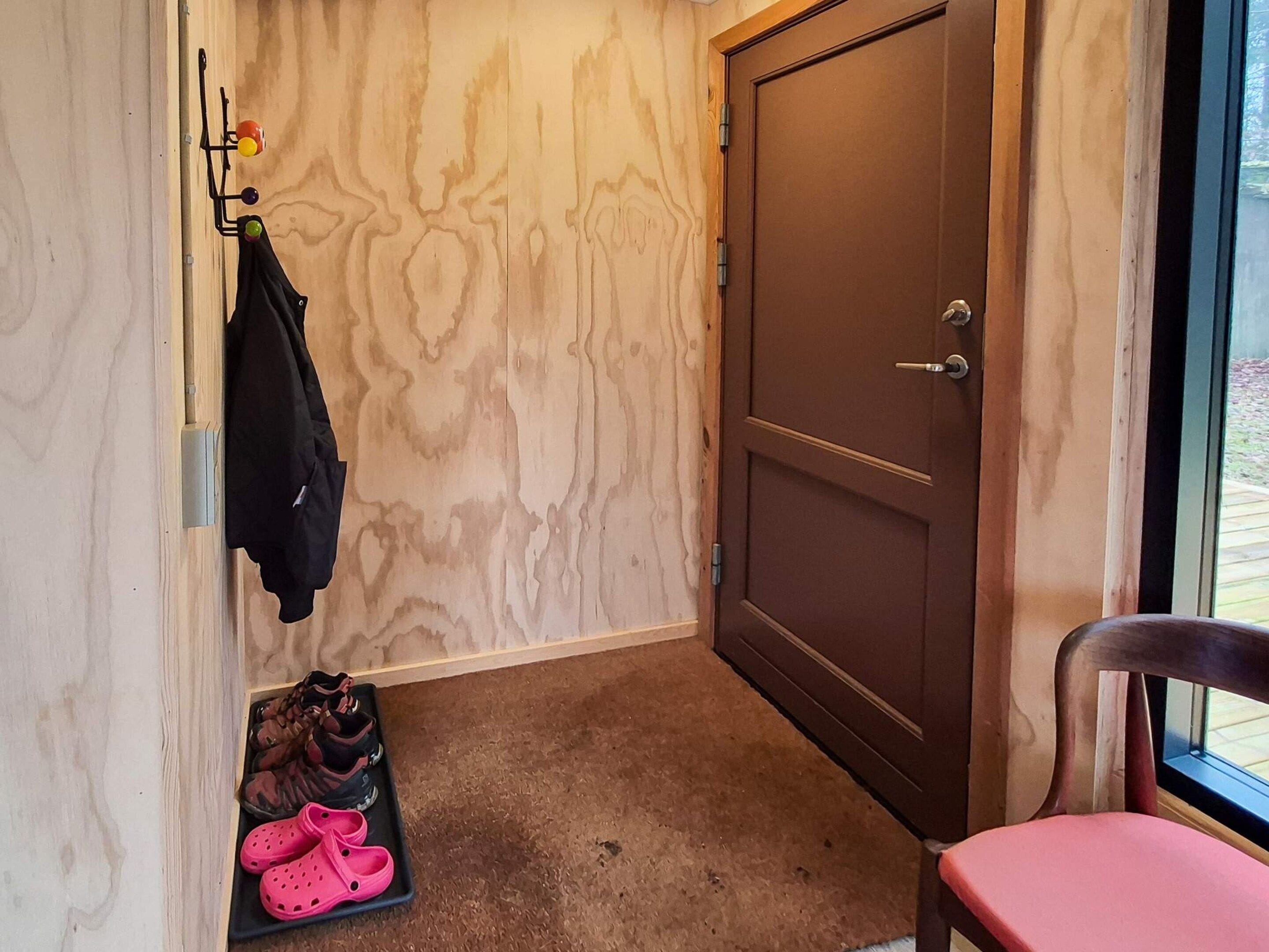 A room with a wooden door, a coat rack with a jacket, a pair of pink slippers, and a chair with a cushion.