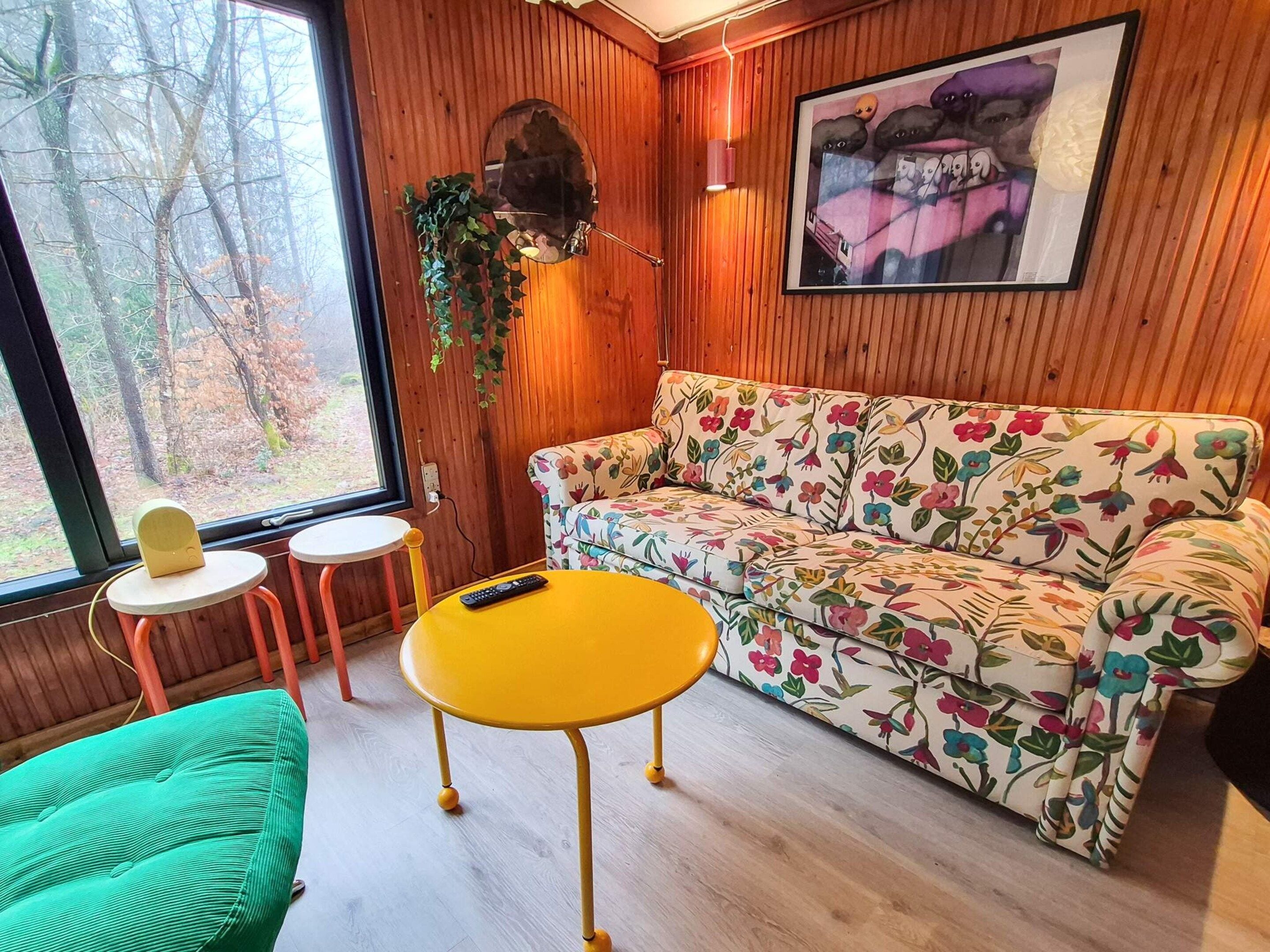 A room with a wooden paneled wall, a floral sofa, a yellow table, a green chair, a small round table, and a framed picture on the wall.