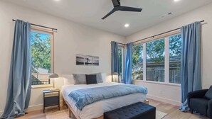 Une chambre à coucher comprenant un lit, une table de chevet, une lampe, un ventilateur de plafond et de grandes fenêtres avec des rideaux.