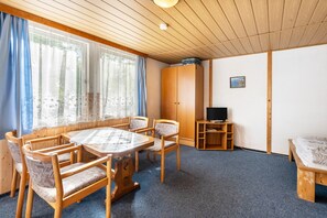 A room with a wooden ceiling, a round table with chairs, a bed, a wardrobe, and a television on a stand.