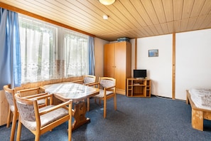 A room with a wooden ceiling, a round table with chairs, a bed, a wardrobe, and a television on a wooden stand.