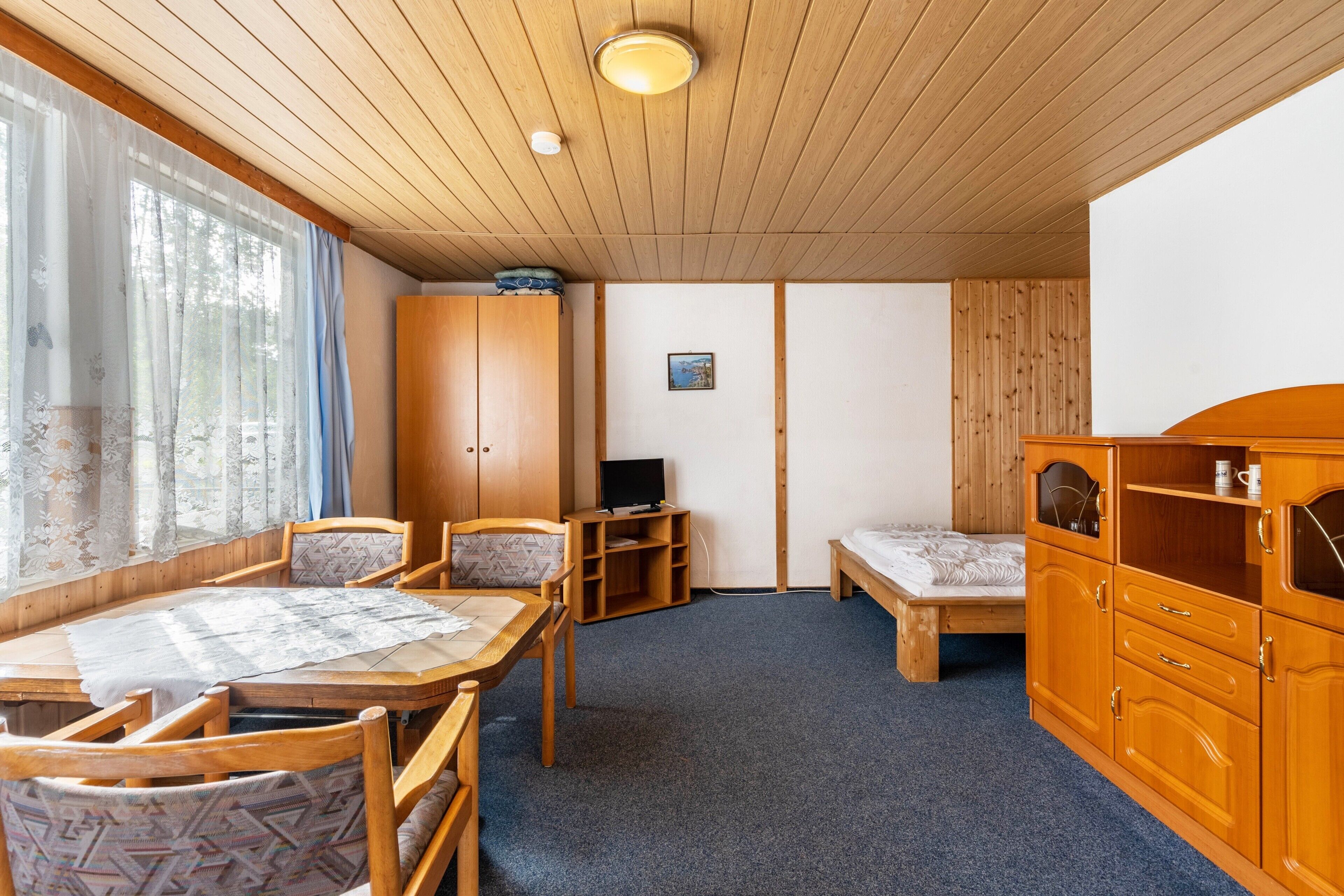 Een kamer met houten plafond, een eettafel met stoelen, een bed, een tv-meubel en een kast.
