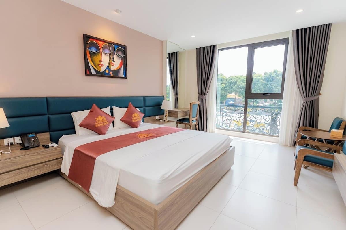 A hotel room with a large bed, a desk with a phone, a chair, a table, and a balcony view.