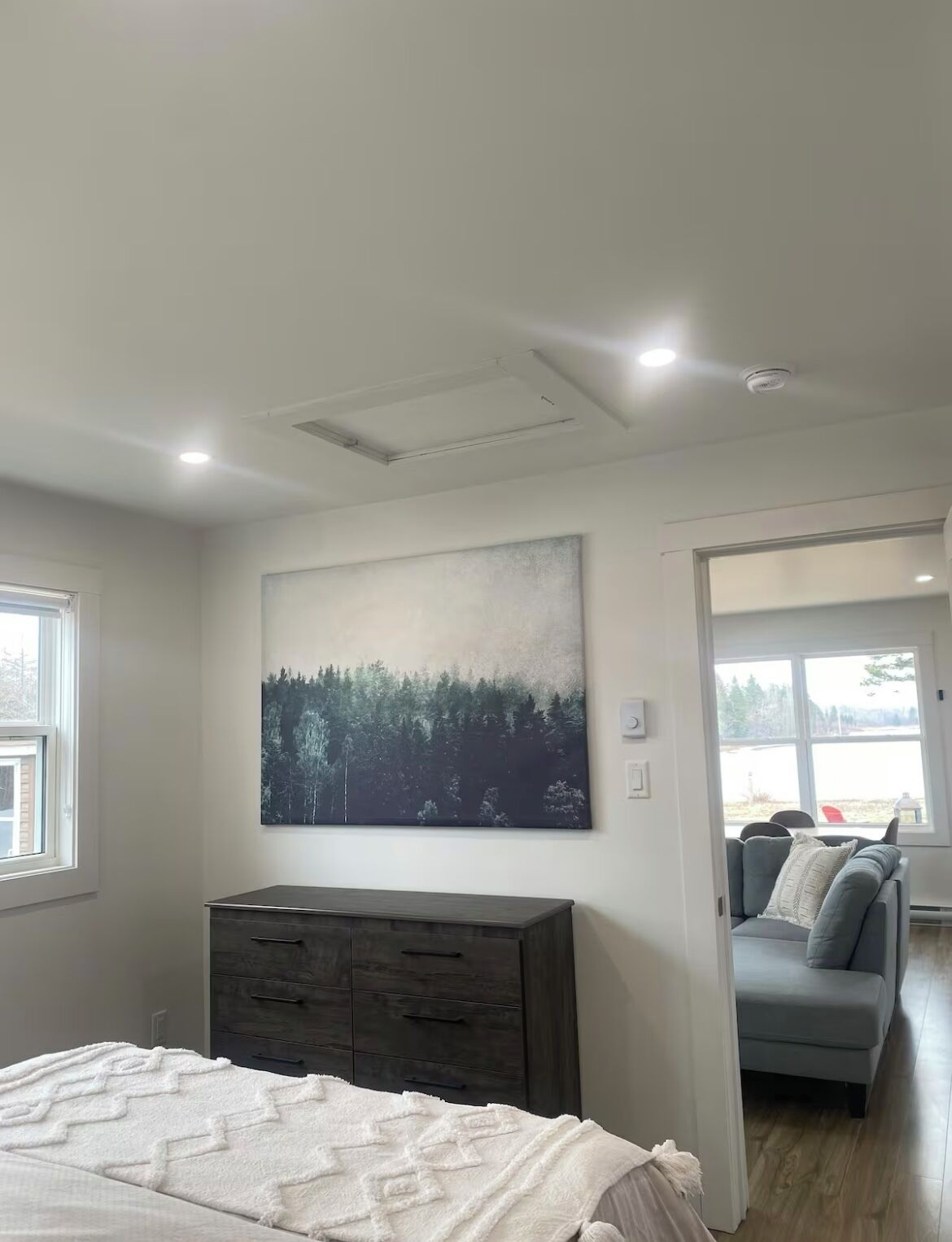 A bedroom with a bed, a dresser, a painting, and a view of a forest through a window.