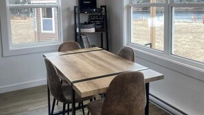A dining area with a wooden table and four chairs, large windows with a view of a park and a building, and a shelf with a sign and a black box. - Waterfront tiny home with hot tub (Pet friendly, near beaches) (Richibucto-Village)