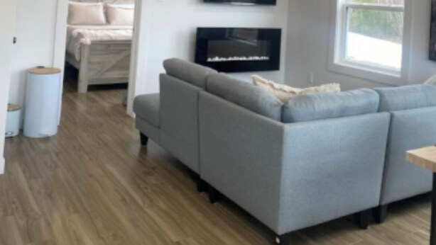 A modern living room with a grey sofa, a flat-screen TV mounted on the wall, and a large window providing natural light.