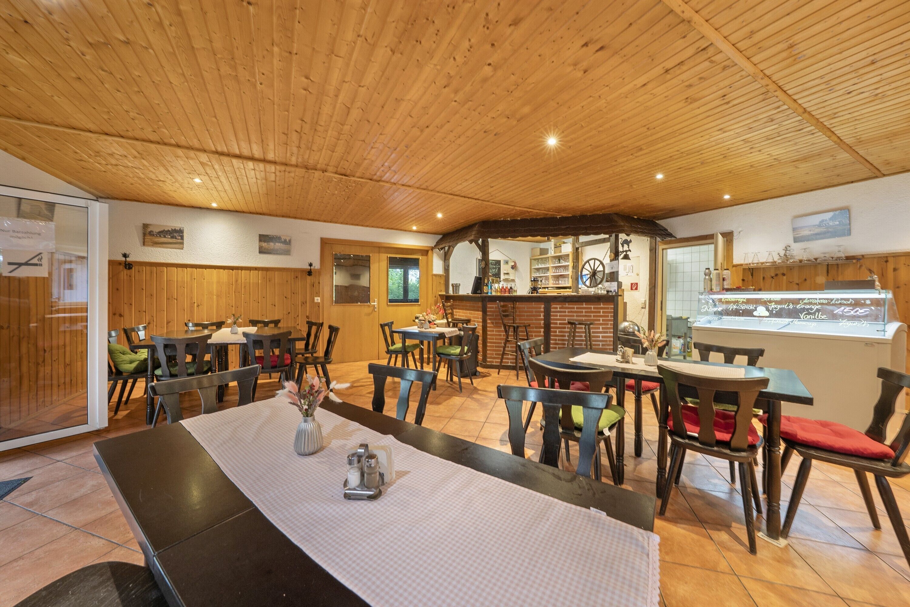 Un restaurante con techo y paredes de madera, mesas con sillas y una barra con vitrina.