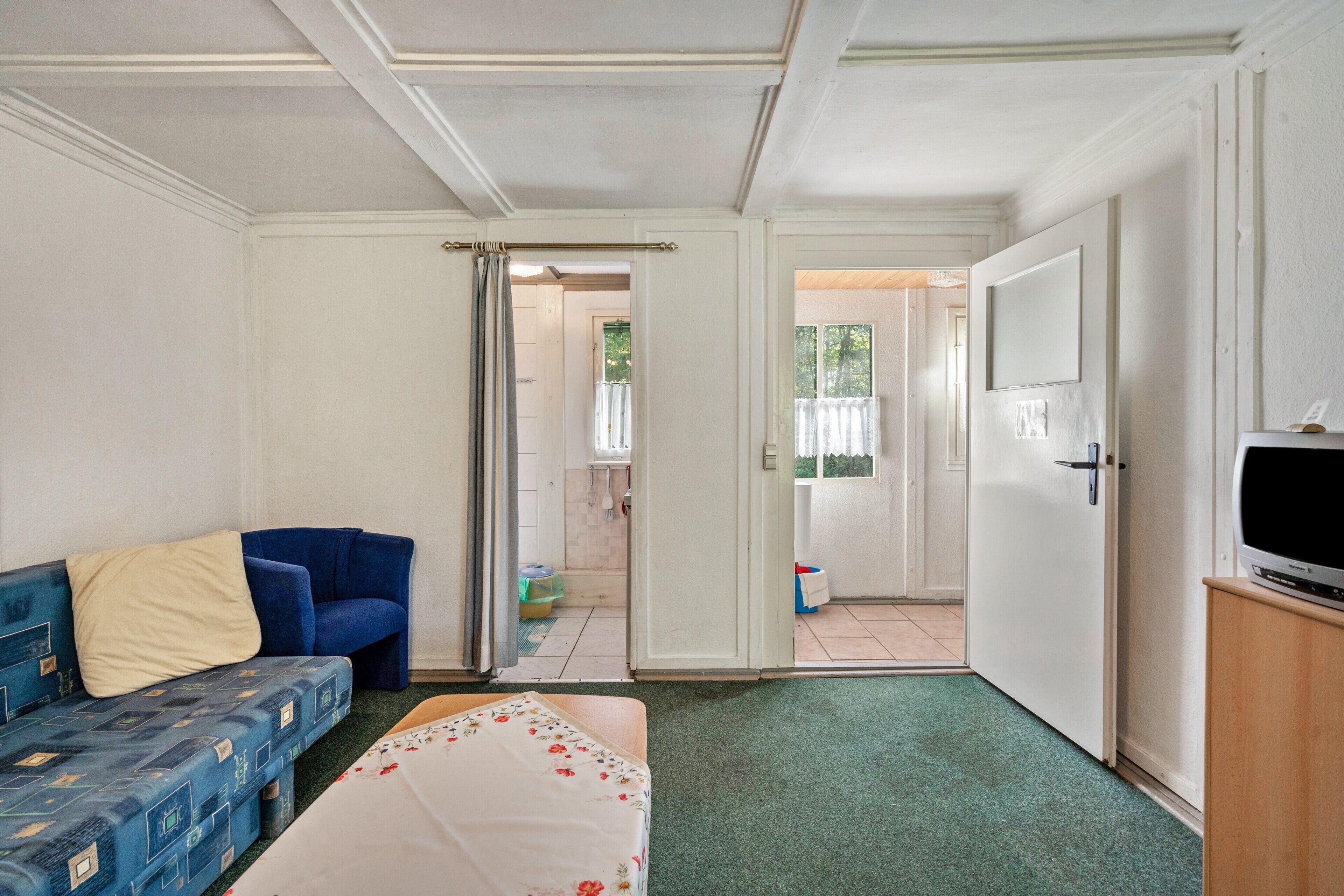 Une pièce avec un canapé bleu, un lit recouvert d’une couvre-lit à motifs floraux, un téléviseur posé sur un meuble en bois et une porte donnant sur un espace extérieur.