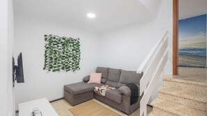 Una sala de estar moderna con un sofá seccional gris, una mesita blanca con adornos, piso de madera y una escalera que lleva a un balcón.