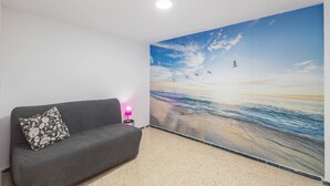 Una habitación con un sofá gris, una lámpara rosa y un mural grande con motivos playeros en la pared.