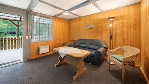 A room with a wooden floor, a table with a white cloth, a leather sofa, a wicker chair, and a wooden wall with a window and a door.