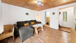 A room with a wooden ceiling, a grey sofa, a wicker chair, a small wooden table, and a door leading to a bathroom.