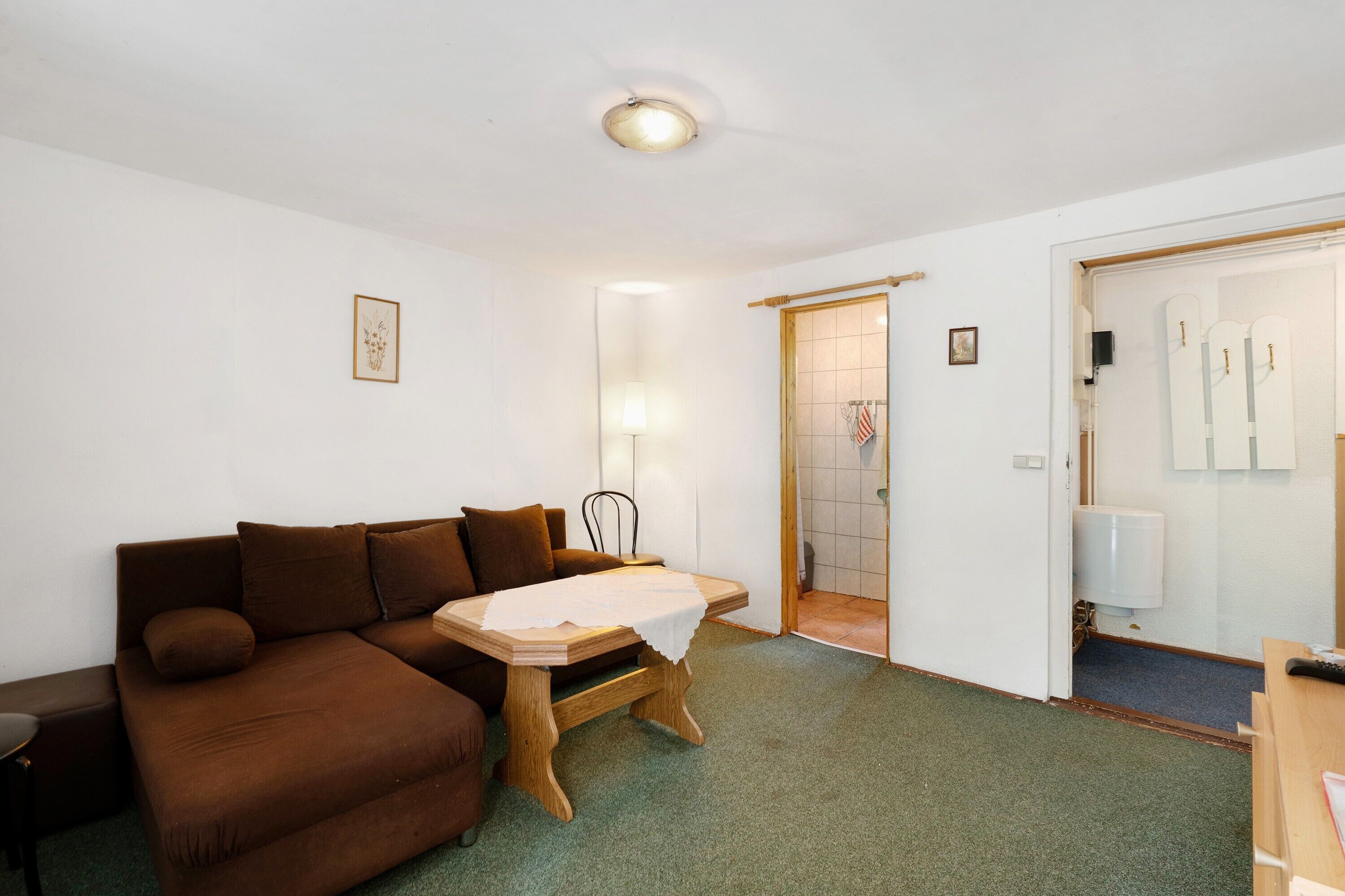 Een kamer met een bruine bank, een houten tafel met een witte tafelkleed, een lamp en een deur naar een badkamer.